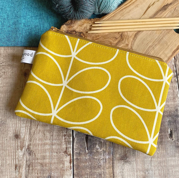A knitting notions pouch sits on a wooden table next to yarn and knitting needles. The pouch is made from very cheerful sunshine yellow fabric and is the perfect size to hold knitting notions. 