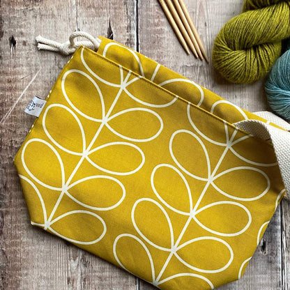 A sunshine yellow knitting project bag lies on a wooden table next to knitting needles and yarn. A woven fabric label is shown with the Eldenwood Craft logo. 