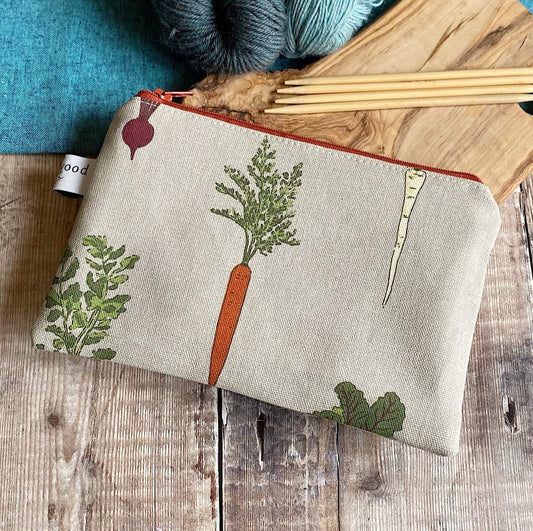 A handmade knitting notions pouch sits on a wooden table next to knitting needles and yarn. The pouch is made from a fabric featuring lovely root vegetables.