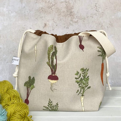 A handmade knitting project bag sits next to some yarn. The bag is covered in a print that features root vegetables including beetroot, carrot, parsnips and turnips. 