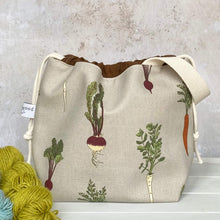 Load image into Gallery viewer, A handmade knitting project bag sits next to some yarn. The bag is covered in a print that features root vegetables including beetroot, carrot, parsnips and turnips. 