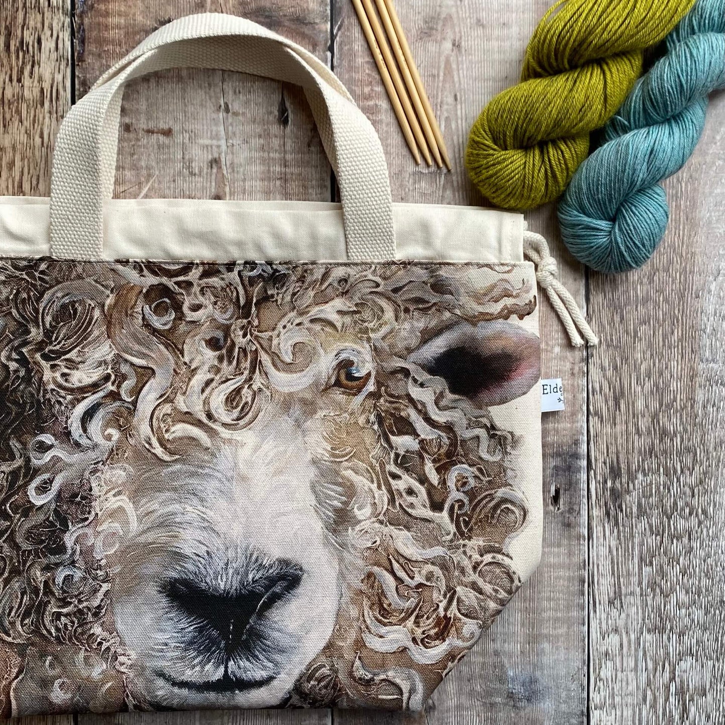 A large knitting project bag lies on a wooden table next to some yarn. From the bag fabric a large sheep face stares out. 