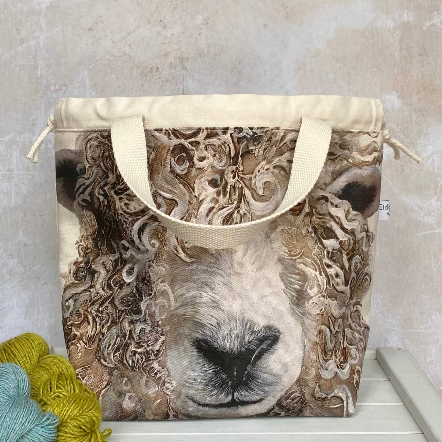 A knitting project bag featuring a large sheep face print sits on a bench. Sturdy natural coloured handles hang down and there is a drawstring closure. 