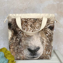 Load image into Gallery viewer, A knitting project bag featuring a large sheep face print sits on a bench. Sturdy natural coloured handles hang down and there is a drawstring closure. 