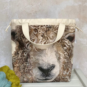 A knitting project bag featuring a large sheep face print sits on a bench. Sturdy natural coloured handles hang down and there is a drawstring closure. 