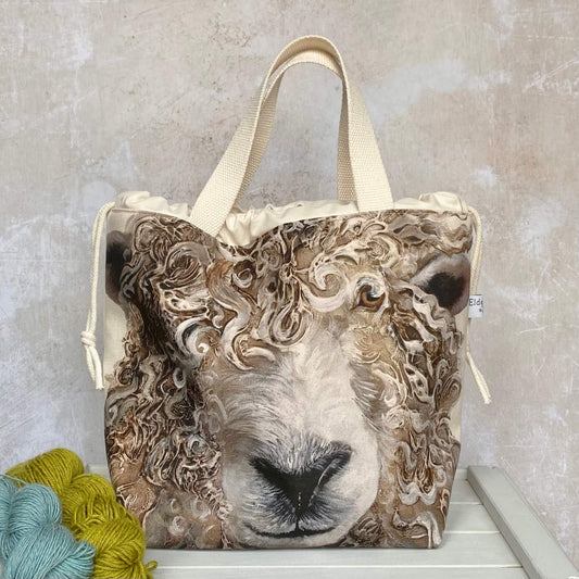 A large knitting project bag sits on a bench next to two skeins of yarn. The bag is handmade and features a long wool sheep face with very curly fleece.