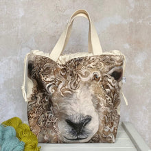 Load image into Gallery viewer, A large knitting project bag sits on a bench next to two skeins of yarn. The bag is handmade and features a long wool sheep face with very curly fleece.