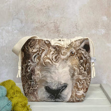Load image into Gallery viewer, A large sheep face adorns the front of a knitting project bag. The bag has a sturdy white handle and sits on a bench next to two skeins of yarn. 