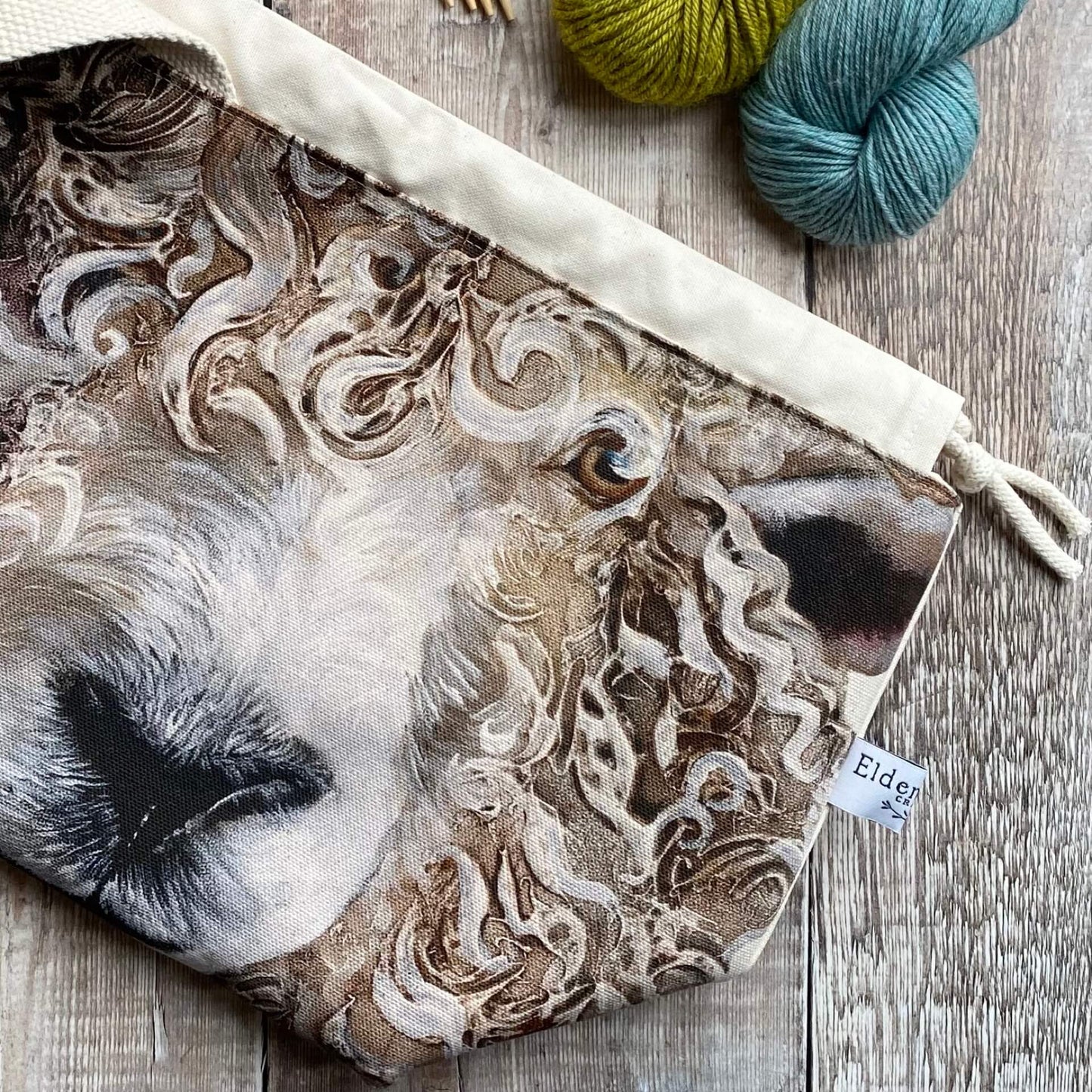 A knitting project bag for medium sized projects lies on a wooden table next to two skeins of yarn. The project bag is handmade and features a large sheep face. 