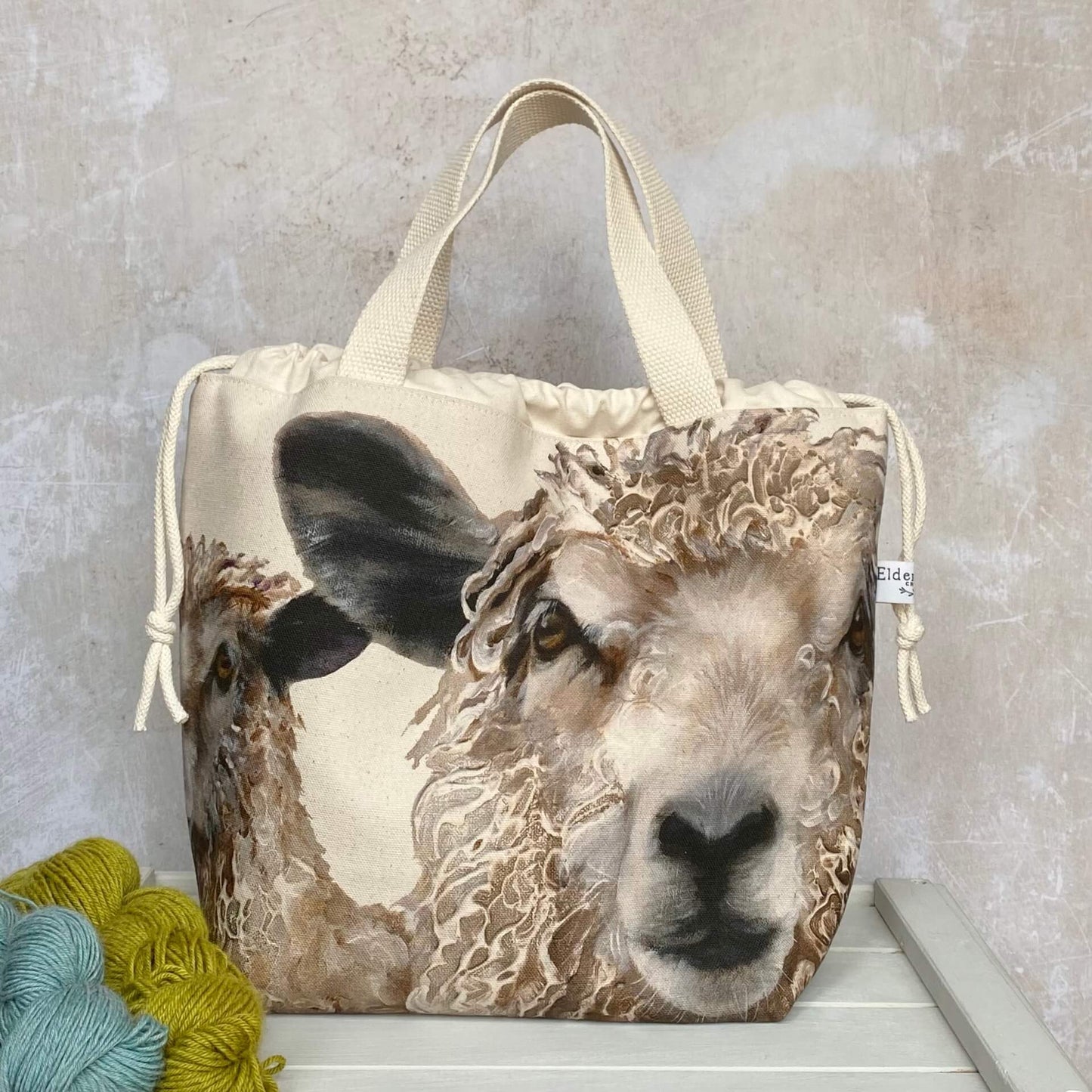 Sheep knitting project bag for large tips. The bag is made from a fabric that features two large sheep faces and is sitting on a bench with two skeins of yarn.