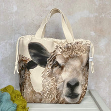 Load image into Gallery viewer, Sheep knitting project bag for large tips. The bag is made from a fabric that features two large sheep faces and is sitting on a bench with two skeins of yarn.
