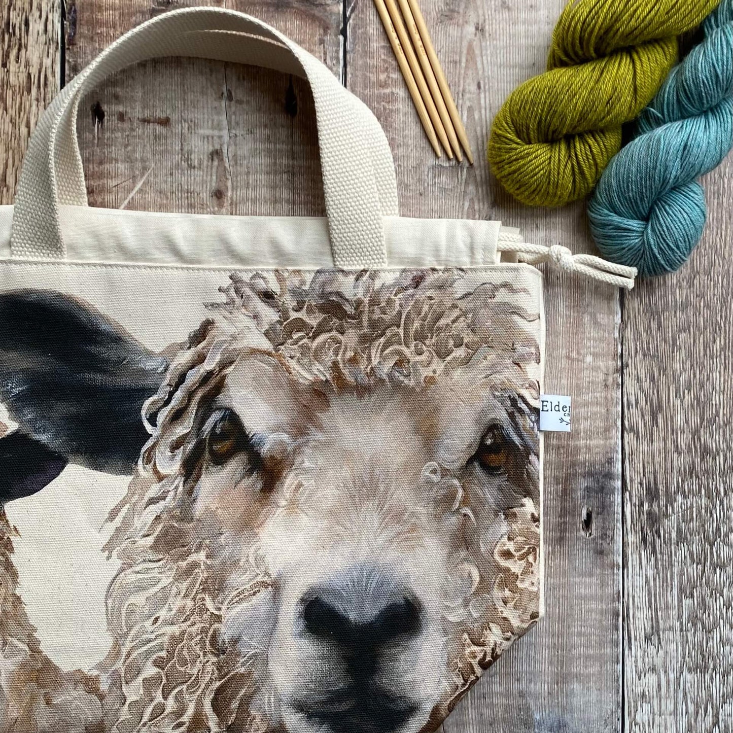A knitting project bag featuring large sheep faces sits on a wooden table next to knitting needles and yarn. The face of one of the sheep stares up at the viewer. 