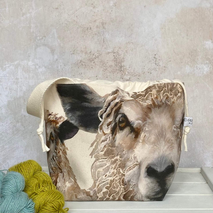 Two sheep stare out of the front of a medium sized knitting project bag that is sitting on top of a bench, next to two skeins of yarn. The project bag has a sturdy natural coloured handle. 
