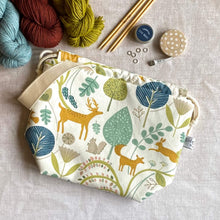 Load image into Gallery viewer, A knitting project bag lies on a table surrounded by yarn and notions. The bag is made from fabric that features Scandi woodland icons such as deer, rabbits, foxes and squirrels as well as lots of trees and leaves. 