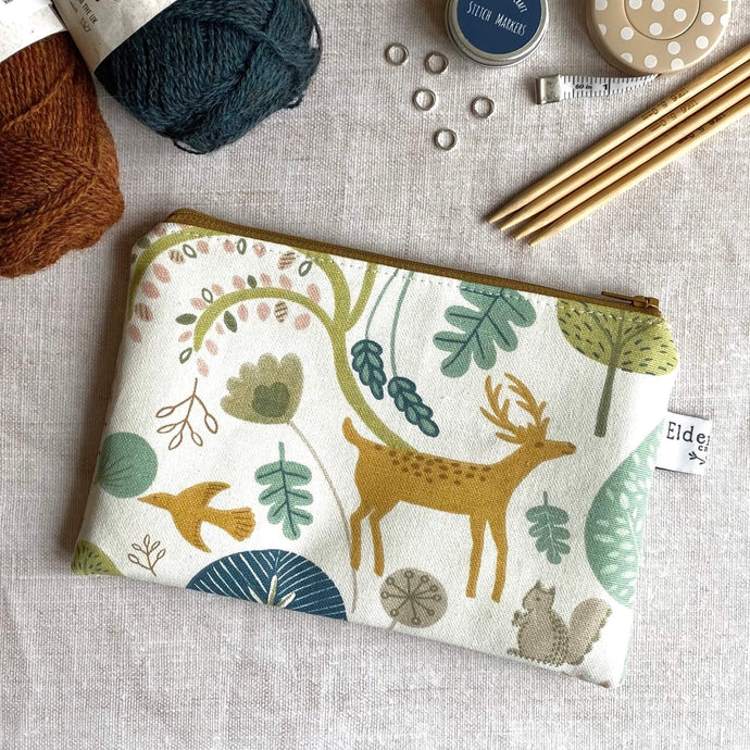 A knitting notions pouch sits on a wooden table. The pouch is hand made from a Scandi woodland print fabric. 