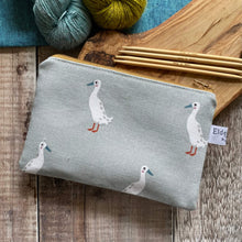 Load image into Gallery viewer, A small zippered pouch for knitting notions sits on a wooden table next to two skeins of yarn, one blue the other green, and some knitting needles. The pouch is made from a soft blue fabric that has some quirky runner ducks printed on it. The pouch has been hand made by Eldenwood Craft. 