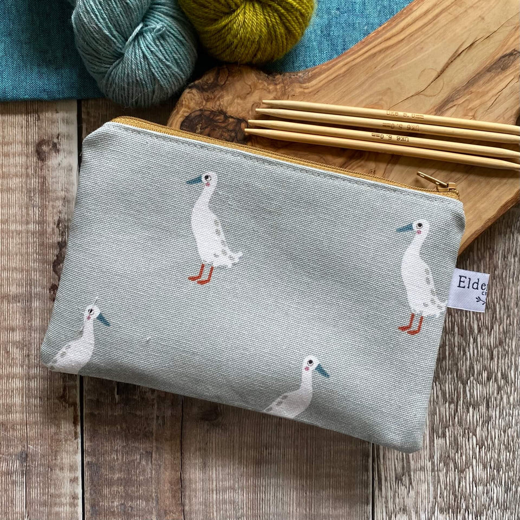 A small zippered pouch for knitting notions sits on a wooden table next to two skeins of yarn, one blue the other green, and some knitting needles. The pouch is made from a soft blue fabric that has some quirky runner ducks printed on it. The pouch has been hand made by Eldenwood Craft. 