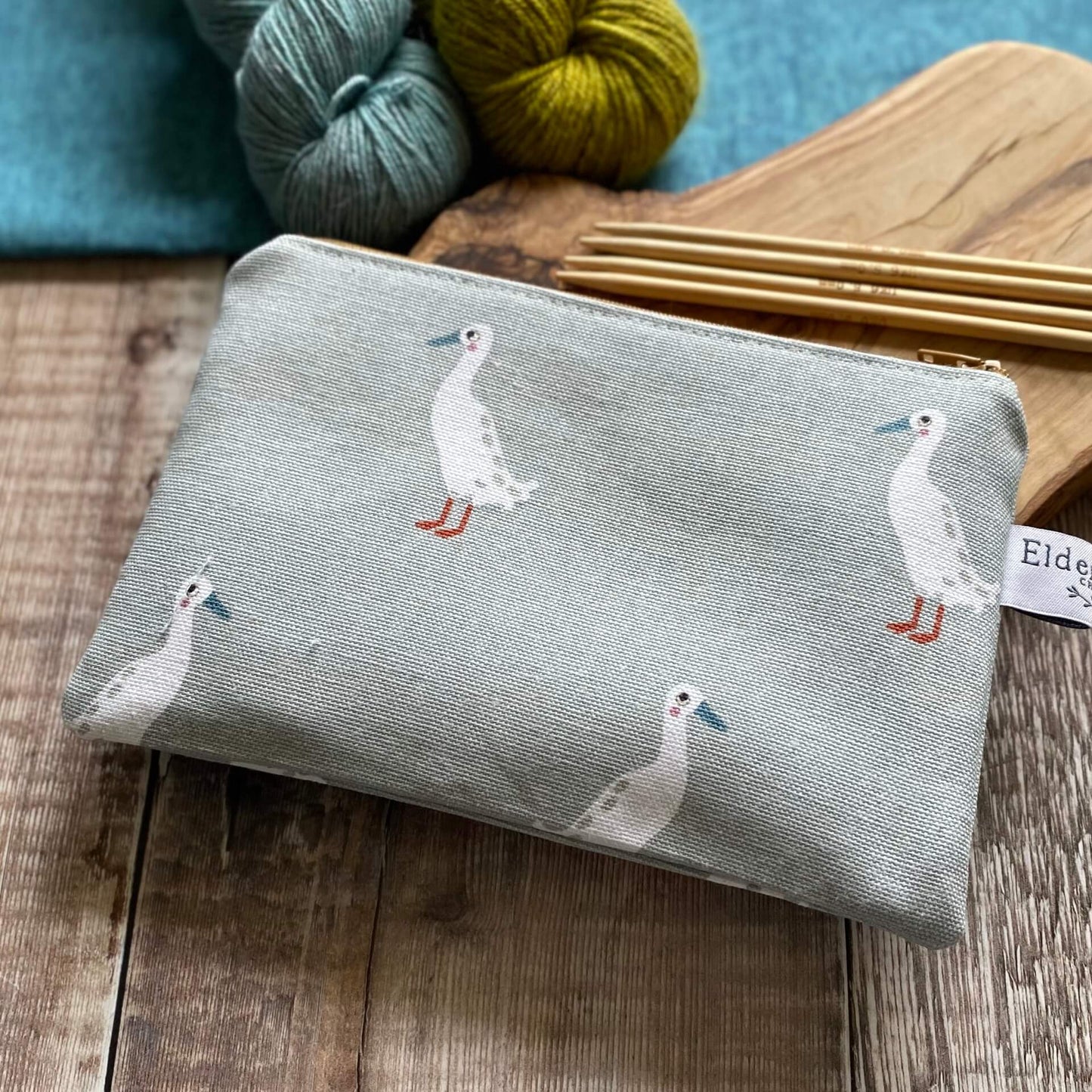 A small zippered pouch for knitting notions, which features some waddling runner ducks, sits on a wooden table next to two skeins of yarn, one blue the other green, and some knitting needles. The pouch has been hand made by Eldenwood Craft. 