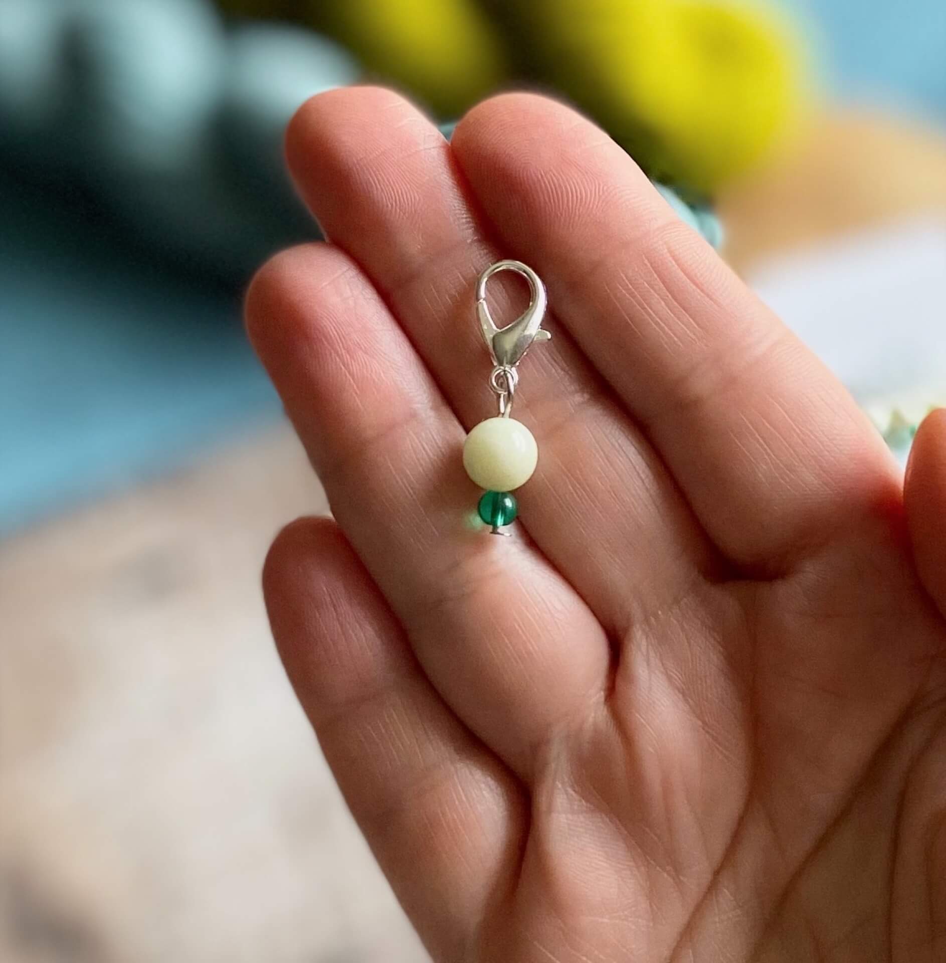 A progress keeper for knitting and crochet, designed to represent a snowdrop, is held on the fingers of the maker's hand. The progress keeper is made up of an ivory bead and a smaller green bead. 