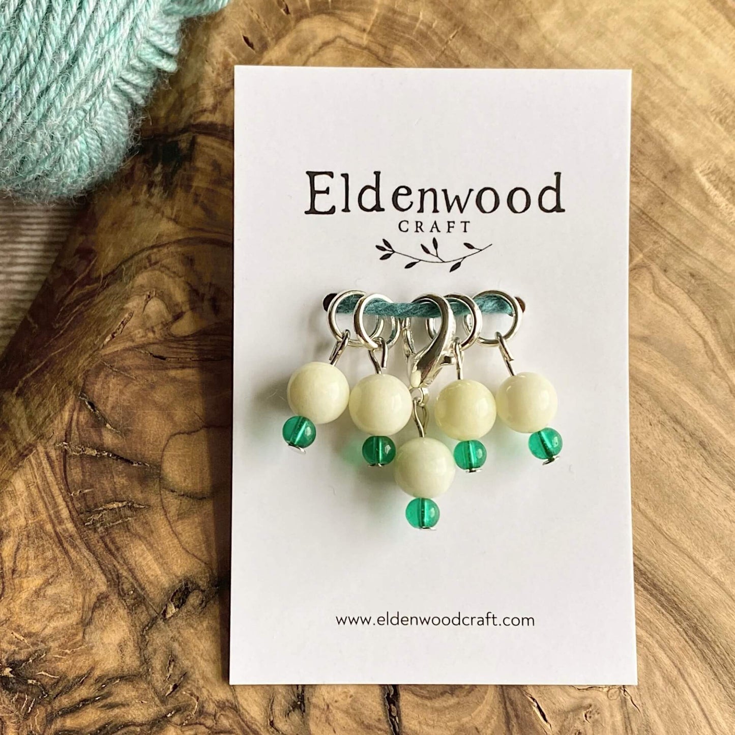A set of four hand made stitch markers and one progress keeper, each designed to represent a snowdrop, sit on a wooden block on a table top, attached to a white Eldenwood Craft card. Just appearing in view is a skein of pale blue yarn. 