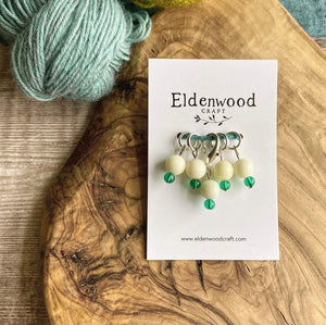A set of four hand made stitch markers and one progress keeper, each designed to represent a snowdrop with an larger ivory  Jasper stone and a small green glass bead, sit on a wooden block on a table top, attached to a white Eldenwood Craft card. 